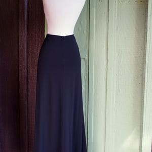 May include: A long, navy blue skirt displayed on a mannequin. The skirt has a white border with small, evenly spaced holes along the bottom edge. The mannequin is white, and the background is a mix of brown and green wooden panels.