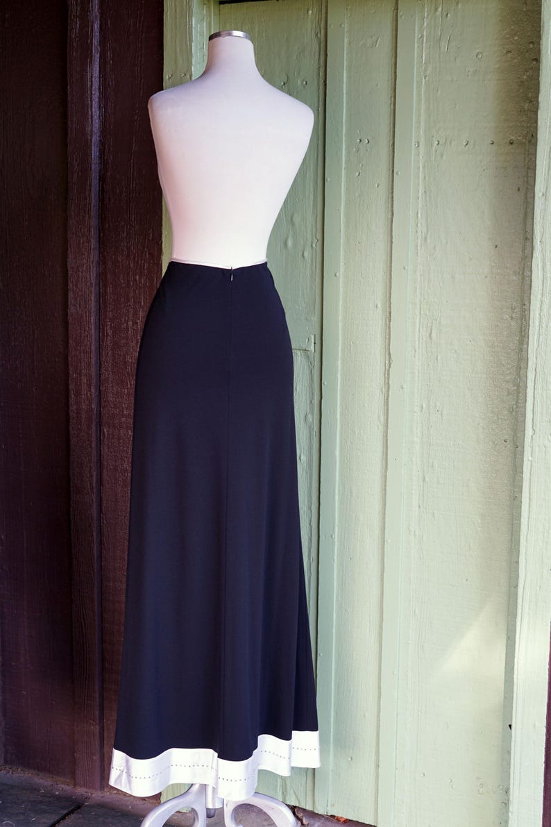 May include: A long, navy blue skirt displayed on a mannequin. The skirt has a white border with small, evenly spaced holes along the bottom edge. The mannequin is white, and the background is a mix of brown and green wooden panels.