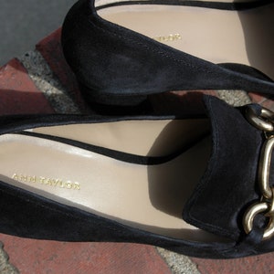 May include: Close-up of two black suede loafers with gold chain detail. The interior of the shoe is a light beige color with the brand name "ANN TAYLOR" printed in gold. The shoes are on a brick surface.