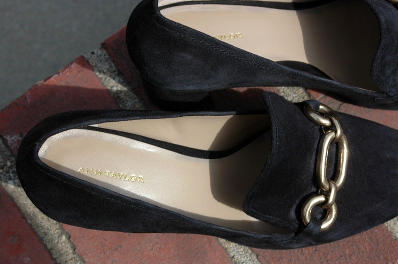 May include: Close-up of two black suede loafers with gold chain detail. The interior of the shoe is a light beige color with the brand name "ANN TAYLOR" printed in gold. The shoes are on a brick surface.