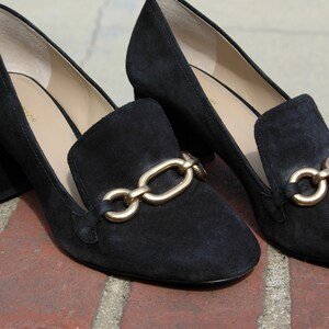 May include: A pair of black suede loafers with a gold chain detail. The shoes have a low block heel and a classic slip-on design. The loafers are displayed on a brick surface.