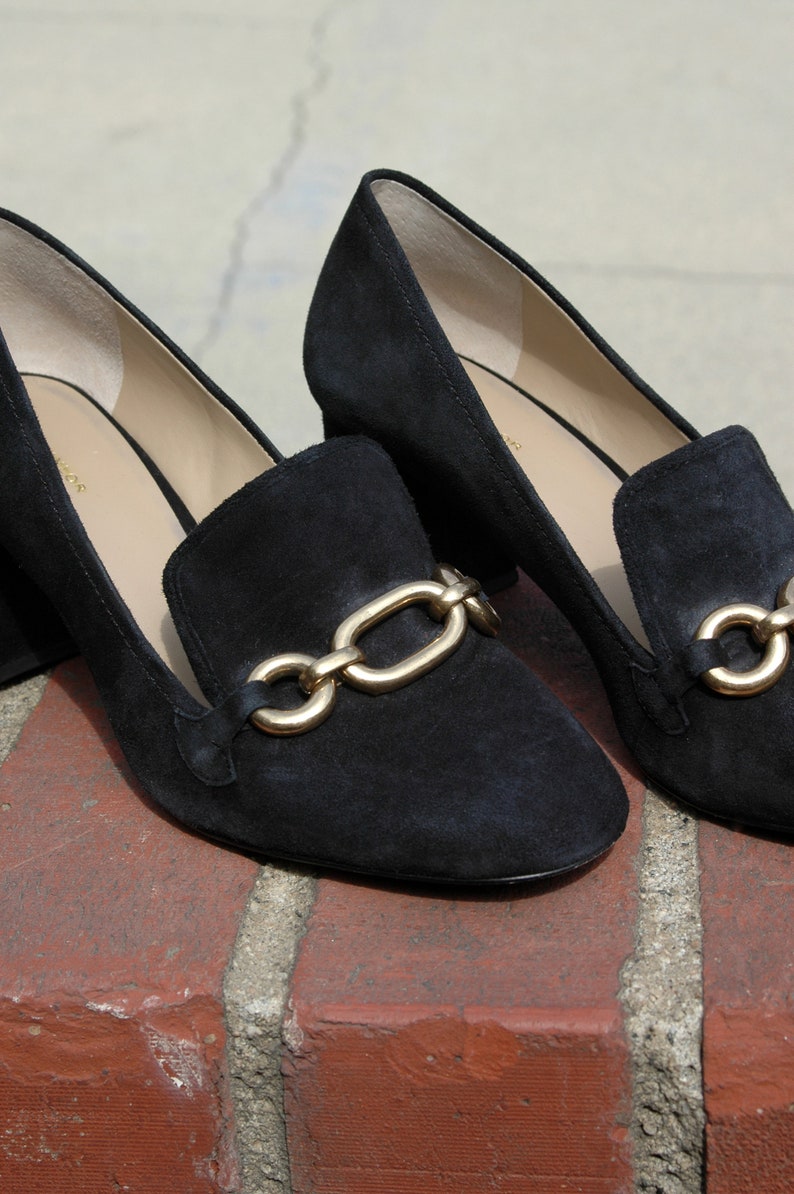 May include: A pair of black suede loafers with a gold chain detail. The shoes have a low block heel and a classic slip-on design. The loafers are displayed on a brick surface.