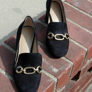 May include: A pair of black suede loafers with gold-tone chain link accents. The shoes are displayed on a red brick surface, showcasing their sleek design and classic style. The loafers have a low heel and a slip-on design.