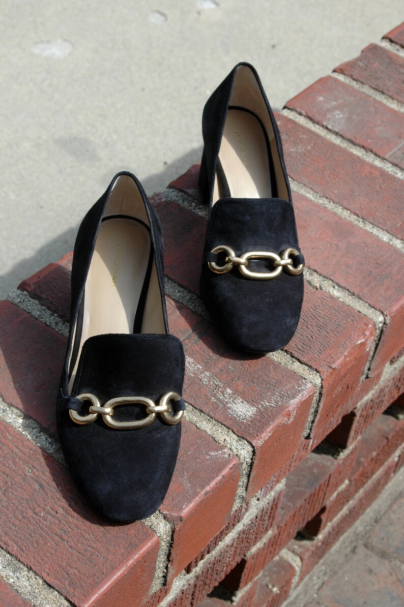 May include: A pair of black suede loafers with gold-tone chain link accents. The shoes are displayed on a red brick surface, showcasing their sleek design and classic style. The loafers have a low heel and a slip-on design.