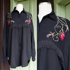 May include: Black button-down shirt with long sleeves, featuring embroidered floral designs in gold, pink, and purple. The shirt has fringe detailing across the chest, adding a unique, vintage flair. The shirt is displayed on a mannequin.