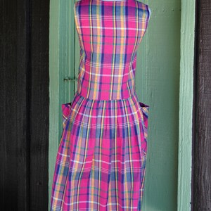 Pu&ograve; includere: Un abito senza maniche a quadri rosa, blu, giallo e bianco con tasche. L'abito &egrave; su un manichino bianco su uno sfondo verde e marrone.
