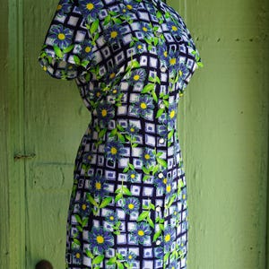 May include: A vintage-style dress on a mannequin. The dress features a floral pattern with blue flowers, green leaves, and yellow centers set against a white and navy blue grid. It has short sleeves and a collared neckline.