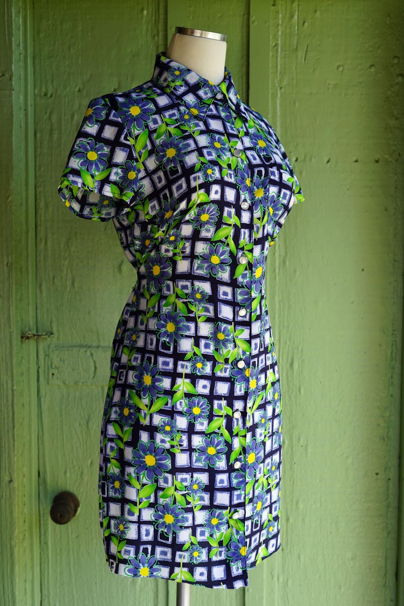 May include: A vintage-style dress on a mannequin. The dress features a floral pattern with blue flowers, green leaves, and yellow centers set against a white and navy blue grid. It has short sleeves and a collared neckline.