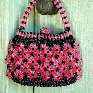 May include: A pink and black beaded purse with a floral design. The purse is hanging on a green door.
