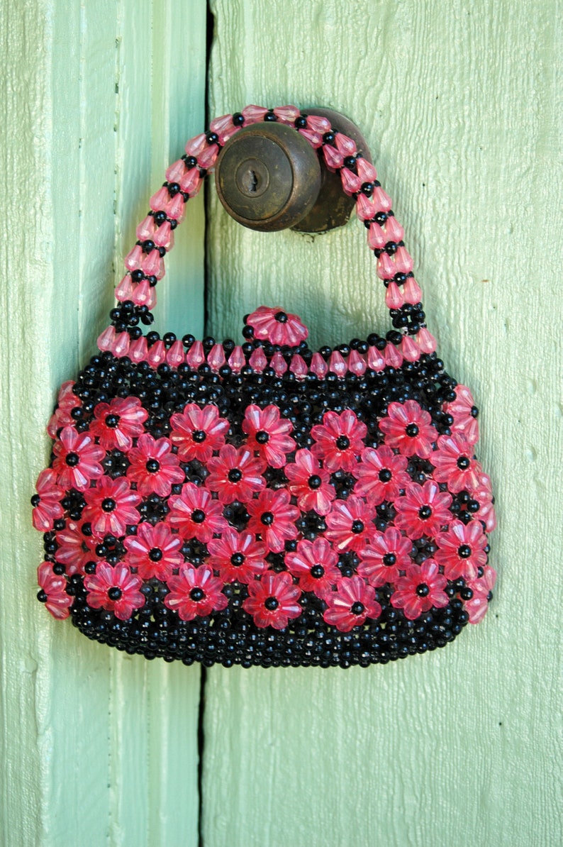 May include: A pink and black beaded purse with a floral design. The purse is hanging on a green door.