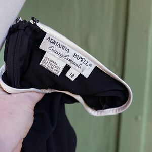 May include: Black evening dress with a white trim and a zipper. The label reads "ADRIANNA PAPELL Evening Essentials" and includes the size 12. The dress is made of rayon and polyester.