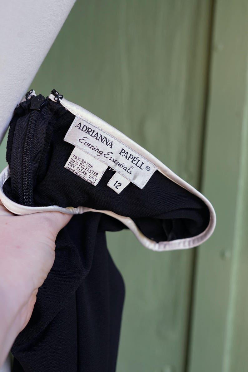 May include: Black evening dress with a white trim and a zipper. The label reads "ADRIANNA PAPELL Evening Essentials" and includes the size 12. The dress is made of rayon and polyester.