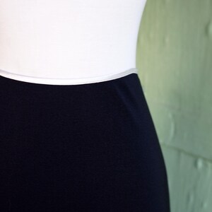 May include: A close-up of a white top and a navy blue skirt. The skirt is a solid color and appears to be made of a smooth fabric. The top is plain and white. The background is a light green wall.