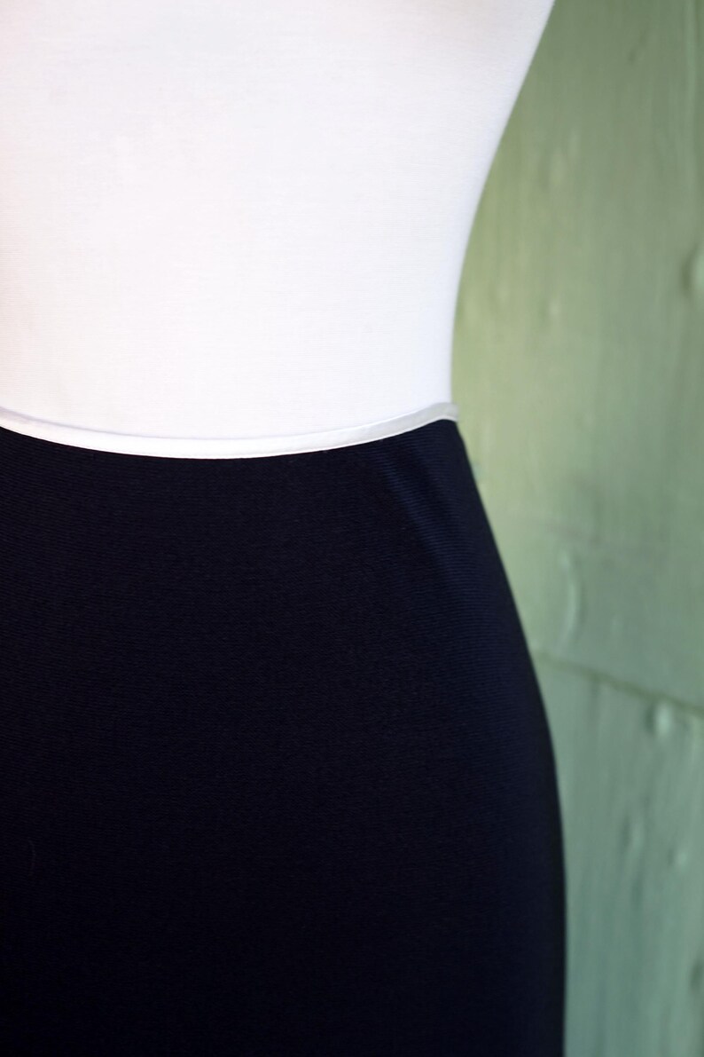 May include: A close-up of a white top and a navy blue skirt. The skirt is a solid color and appears to be made of a smooth fabric. The top is plain and white. The background is a light green wall.