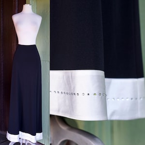 May include: A black maxi skirt with a white band at the hem, embellished with small, sparkling accents. The skirt is displayed on a mannequin, showcasing its elegant, flowing design. The skirt's length is approximately 40 inches.