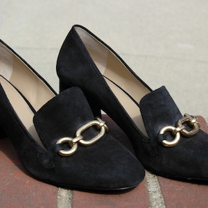 May include: A pair of black suede heeled loafers with gold-tone chain detailing. The shoes have a block heel and a classic loafer design, suitable for various occasions.