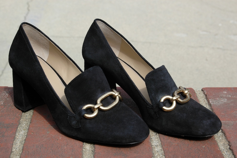 May include: A pair of black suede heeled loafers with gold-tone chain detailing. The shoes have a block heel and a classic loafer design, suitable for various occasions.