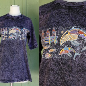 May include: Purple tie-dye t-shirt with a colorful graphic of dolphins, fish, and people in a canoe. The graphic is in a style reminiscent of Aboriginal art.