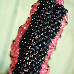 May include: A black and pink beaded necklace with a unique woven design. The beads are small and round, and the necklace is long enough to be worn as a choker or a longer necklace.