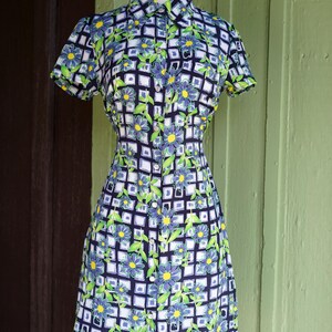 May include: A short-sleeved dress with a floral pattern. The dress features a collared neckline and a button-down front. The pattern includes blue flowers, green leaves, and a white grid design on a navy blue background. The dress is displayed on a mannequin.