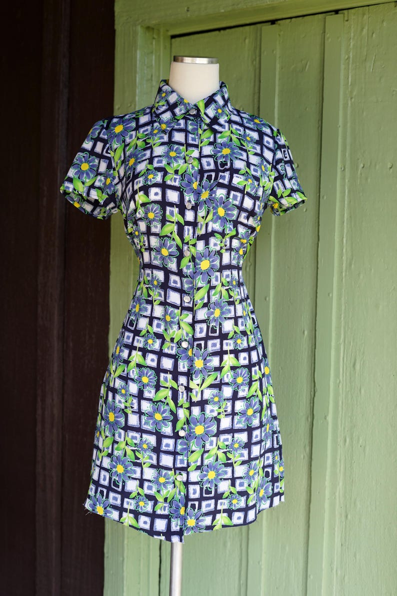 May include: A short-sleeved dress with a floral pattern. The dress features a collared neckline and a button-down front. The pattern includes blue flowers, green leaves, and a white grid design on a navy blue background. The dress is displayed on a mannequin.