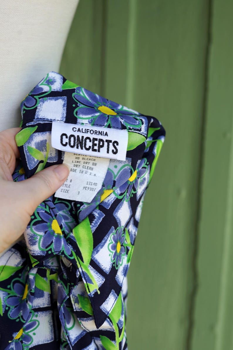 May include: A close-up of a patterned shirt with a floral design in blue, green, and yellow on a navy blue background. The shirt has a white tag that reads "CALIFORNIA CONCEPTS". The shirt is size 3 and made in the U.S.A.