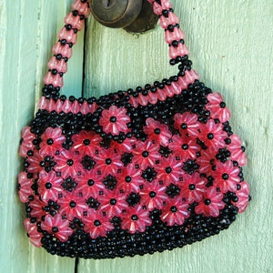 May include: A small pink and black beaded purse with a flower design. The purse is hanging on a green door.