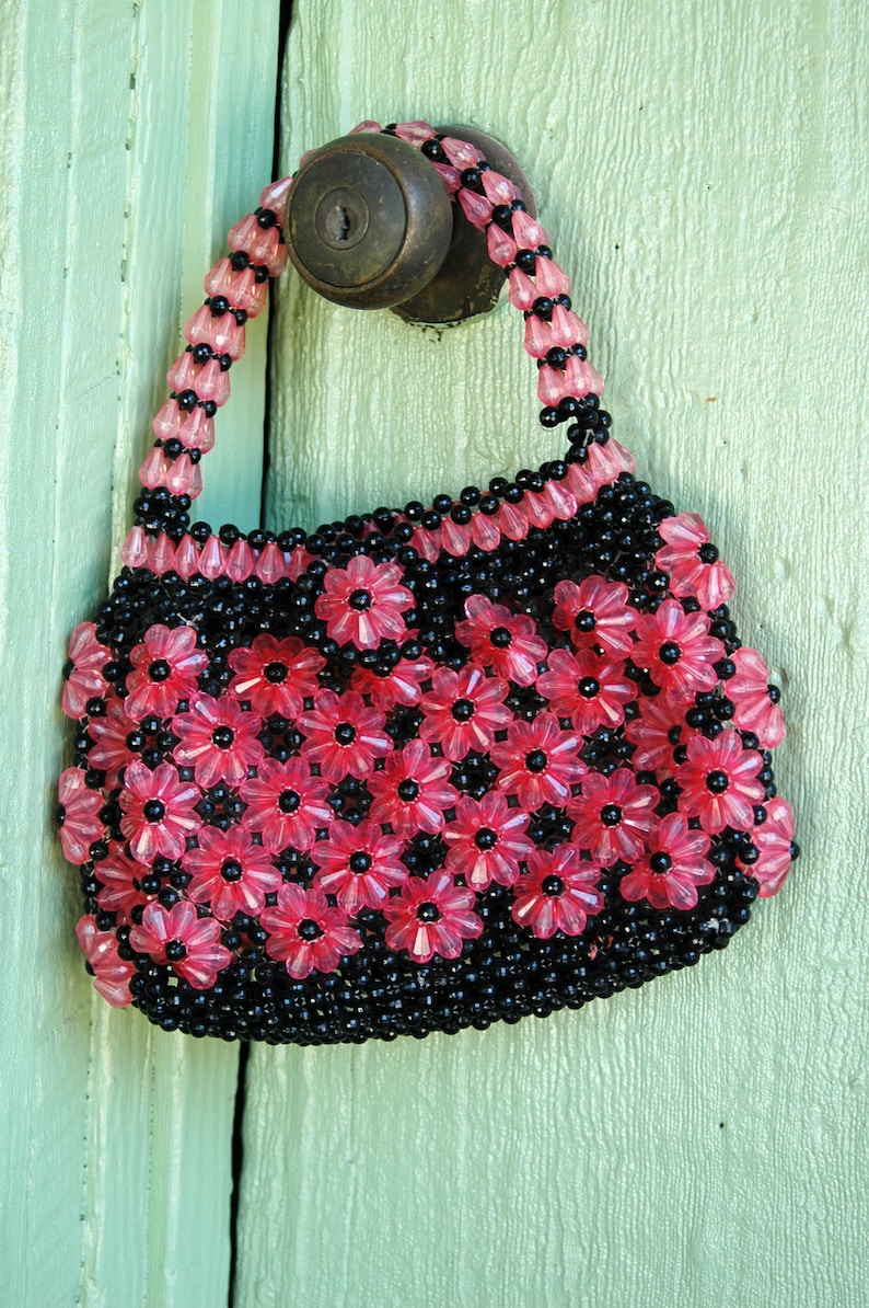 May include: A small pink and black beaded purse with a flower design. The purse is hanging on a green door.