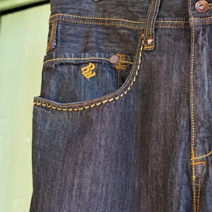 Peut inclure: Un jean en denim fonc&eacute; avec des d&eacute;tails de couture orange contrast&eacute;s. Le jean a une poche avant avec un petit logo au centre.