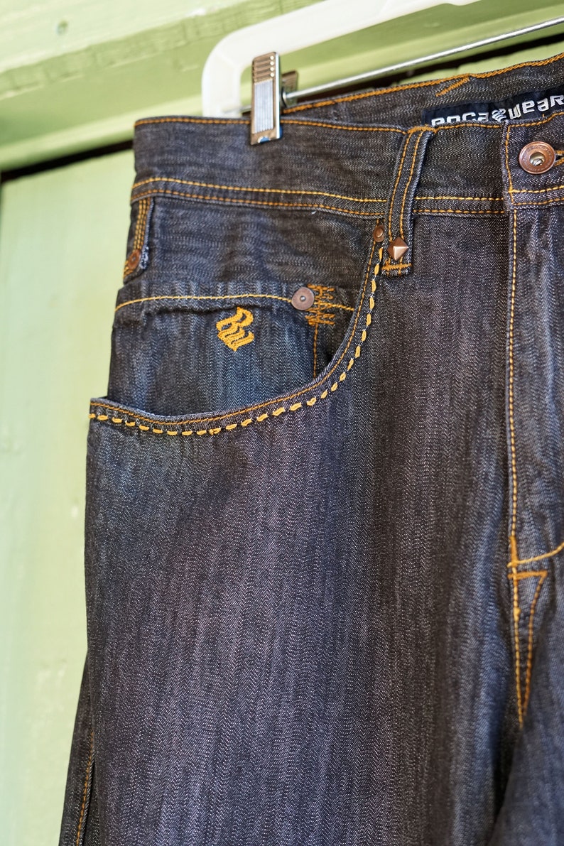 Peut inclure: Un jean en denim fonc&eacute; avec des d&eacute;tails de couture orange contrast&eacute;s. Le jean a une poche avant avec un petit logo au centre.