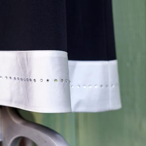May include: Close-up of two black fabric panels with white satin trim. The trim is embellished with a row of small, sparkling rhinestones. The fabric appears to be part of a garment, possibly a skirt or dress.