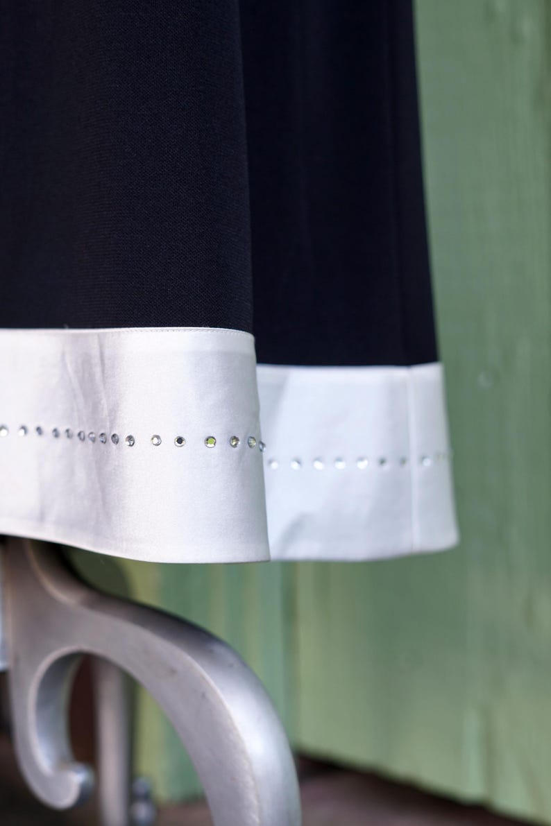 May include: Close-up of two black fabric panels with white satin trim. The trim is embellished with a row of small, sparkling rhinestones. The fabric appears to be part of a garment, possibly a skirt or dress.