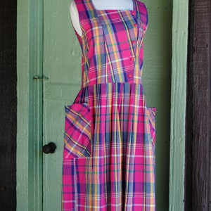 Pu&ograve; includere: Un abito senza maniche a quadri rosa, blu e giallo con scollo quadrato e tasche. L'abito &egrave; su un manichino bianco su uno sfondo di legno verde.