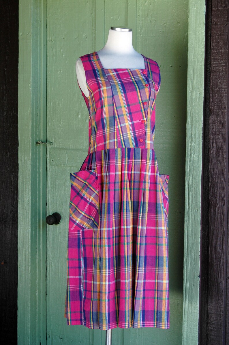 Pu&ograve; includere: Un abito senza maniche a quadri rosa, blu e giallo con scollo quadrato e tasche. L'abito &egrave; su un manichino bianco su uno sfondo di legno verde.