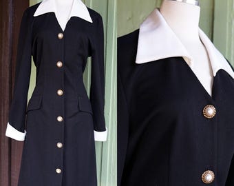 Puede incluir: Un abrigo-vestido negro con cuello y puños blancos. El abrigo tiene botones dorados y dos bolsillos delanteros. El vestido se exhibe en un maniquí.