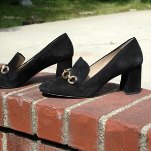 May include: A pair of black suede heeled loafers with gold-colored chain accents. The shoes have a block heel and are displayed on a red brick wall, with a blurred outdoor background.