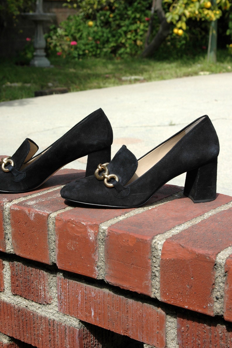 May include: A pair of black suede heeled loafers with gold-colored chain accents. The shoes have a block heel and are displayed on a red brick wall, with a blurred outdoor background.
