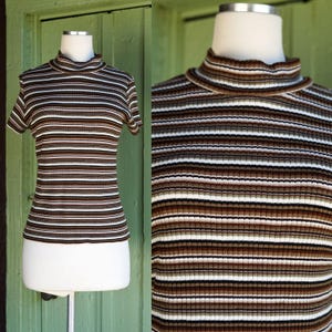 May include: A short-sleeved, ribbed knit top with a mock turtleneck. The top features horizontal stripes in shades of brown, white, and black. The garment is displayed on a mannequin against a green background.