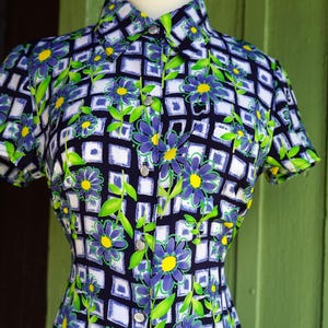 May include: A vintage button-down shirt with a floral pattern. The shirt features a navy blue background with a grid of white squares, adorned with purple flowers, yellow centers, and green leaves. The short-sleeved shirt is displayed on a mannequin.