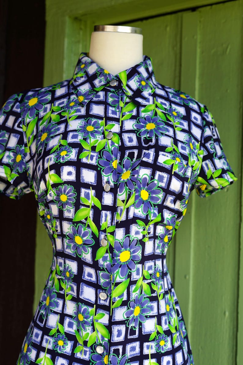 May include: A vintage button-down shirt with a floral pattern. The shirt features a navy blue background with a grid of white squares, adorned with purple flowers, yellow centers, and green leaves. The short-sleeved shirt is displayed on a mannequin.
