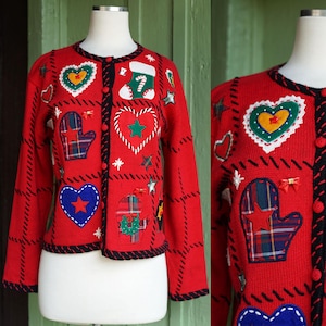 May include: A red cardigan sweater with Christmas-themed appliqués. The sweater features a black trim, button closure, and various patches including hearts, mittens, stars, and a stocking. The appliqués are in various colors, including green, blue, and yellow.