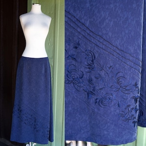 Puede incluir: Una falda azul marino con un diseño floral. La falda se muestra en un maniquí. La falda tiene un patrón sutil y un bordado floral detallado a lo largo del dobladillo y una raya diagonal.