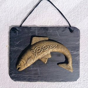 May include: A bronze fish sculpture with Celtic knot detailing, mounted on a dark grey slate plaque. The fish has detailed scales and fins. A black cord is attached for hanging. The plaque is rectangular.
