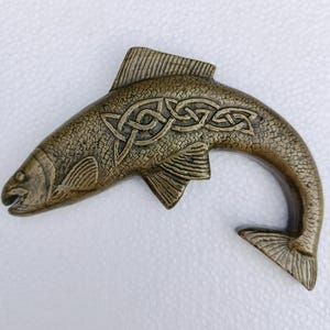 Puede incluir: Objeto decorativo en forma de pez de color bronce con diseño de nudo celta. El pez tiene escamas y aletas detalladas, y una cola curvada. El objeto es probablemente de metal y tiene una apariencia antigua o vintage.