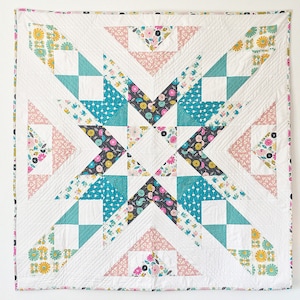 May include: A white quilt with a geometric pattern in shades of teal, pink, and gray. The quilt is made up of triangles and squares, and the pattern is centered around a large star shape.