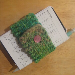 May include: A green and brown knitted wrist warmer with a pink button. The wrist warmer is covering a small white card with the text "Morse Code" and a chart of Morse code.
