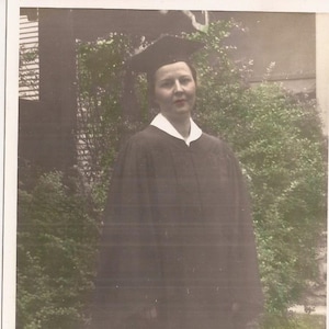 May include: A woman in a black graduation gown and cap, standing in front of a green bush.