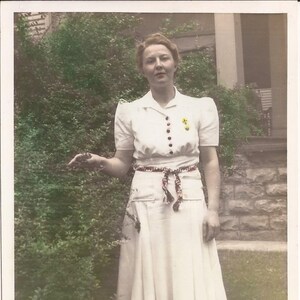 May include: A woman in a white dress with a red and white checkered sash stands in front of a green bush. She is smiling and looking at the camera.