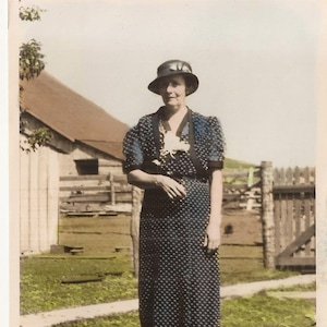 May include: A woman in a black and white polka dot dress and a black hat stands in front of a wooden fence and a building. She is smiling and looking at the camera. There are pink flowers in the foreground.