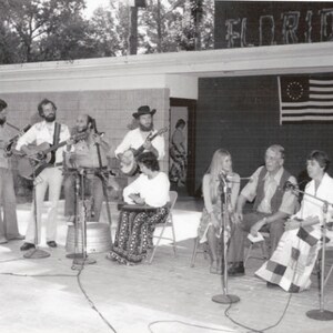 May include: A group of people playing music outdoors. They are standing on a stage with a sign that says "FLORIDA" above them. The people are playing guitars and other instruments. There is a microphone in front of each person.